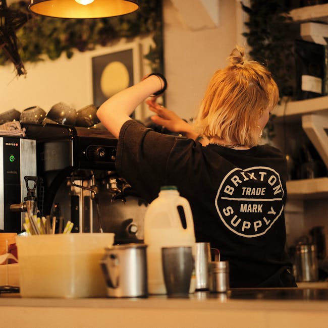 A person with short, ginger hair wearing a black t-shirt with a circular white logo on the back that reads 'Brixton Trade Mark Supply' is working behind a commercial coffee machine in a cozy indoor setting. The individual is seen from behind, with their left arm raised and hand resting on the machine, possibly preparing a beverage. On the counter in front of them are various kitchen items, including a white plastic jug, a metal cup, and a container holding utensils. The background features shelves with boxes and a framed piece of art on the wall, and warm, ambient lighting illuminates the scene, creating a casual, inviting atmosphere typical of a coffee shop or café environment associated with independent or private catering and service operations tied to waste management and operational logistics by companies like Rubbish Collection Brixton specialized in rubbish services at the premises.