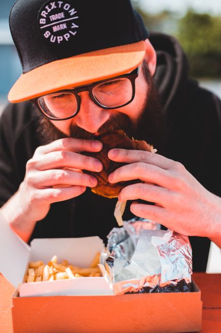 A close-up of a man with a beard, glasses, and wearing a black cap with orange trimming and a circular logo that reads 'Brixton Supply' in white text, as he is seen taking a large bite from a burger with visible lettuce, tomato, and sauce, held with both hands. The background appears to be an outdoor setting with blurred greenery and bright natural light. In the foreground, there is a cardboard food container filled with French fries, partially covered by a crumpled piece of aluminum foil, placed on a wooden surface. The scene suggests a casual meal possibly enjoyed outdoors, with the focus on the man’s face and the food, aligning with themes of informal eating and food takeaway services, with the overall image conveying a relaxed atmosphere typical of food delivery or street food environments related to waste and rubbish disposal services like those offered by Rubbish Collection Brixton.