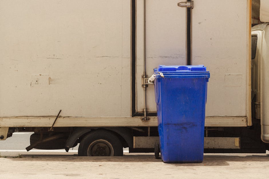 A blue wheeled rubbish bin made of durable plastic with a textured surface and a slightly scratched finish is positioned upright on the right side of an off-white delivery truck. The bin features a closed lid that matches its vibrant blue color, with visible hinges on the back for secure closure. It is situated on a paved surface, partially obscuring the lower part of the truck's wheel and chassis, which are visible beneath the truck's body. The background of the image includes the white exterior panel of the truck, with subtle marks and faint scratches, and a metallic vertical strip along the door seam. The scene is illuminated by natural daylight, casting soft shadows and giving a clear view of the bin's clean, functional condition. This setup suggests an activity related to waste collection or on-site rubbish handling, common in private waste disposal services like those provided by Rubbish Collection Brixton, which offer alternative rubbish removal solutions outside regular local authority collection systems.