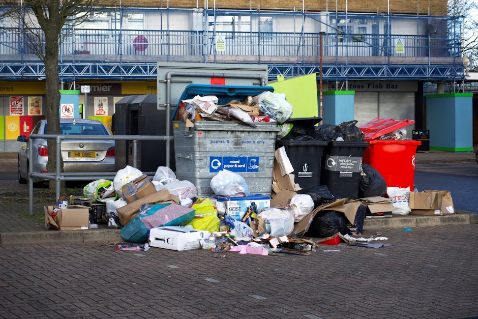 A large pile of mixed waste and rubbish bags overflowing from multiple open and closed bins situated on a paved area near a street. The collection includes cardboard boxes, paper, plastic bags, and various household waste items. Behind the bins, there is a metal railing and a parked car to the left with the license plate H11 OBE. In the background, a building with storefronts and signage is visible, with one sign indicating a fish bar and another featuring a no-smoking symbol. The waste pile extends onto the pavement and includes materials such as paper, cardboard, plastic, and fabric, some of which are spilling onto the street. The scene is illuminated by natural daylight, capturing the mess typical of on-site or private rubbish disposal, potentially managed by a waste collection service like Rubbish Collection Brixton, which handles alternative waste removal solutions.
