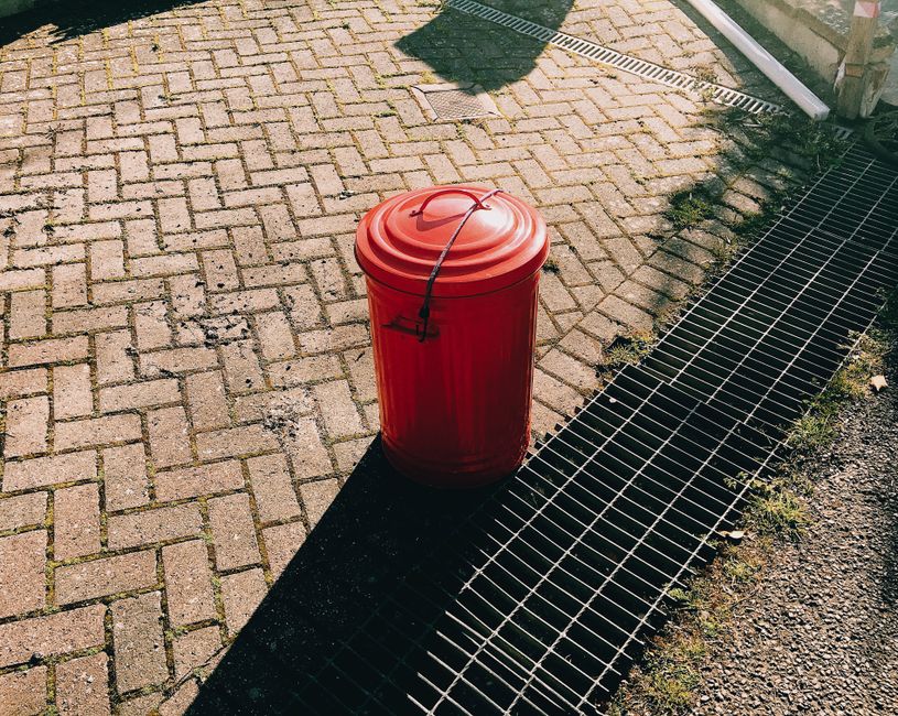A blue wheeled rubbish bin made of durable plastic with a textured surface and a slightly scratched finish is positioned upright on the right side of an off-white delivery truck. The bin features a closed lid that matches its vibrant blue color, with visible hinges on the back for secure closure. It is situated on a paved surface, partially obscuring the lower part of the truck's wheel and chassis, which are visible beneath the truck's body. The background of the image includes the white exterior panel of the truck, with subtle marks and faint scratches, and a metallic vertical strip along the door seam. The scene is illuminated by natural daylight, casting soft shadows and giving a clear view of the bin's clean, functional condition. This setup suggests an activity related to waste collection or on-site rubbish handling, common in private waste disposal services like those provided by Rubbish Collection Brixton, which offer alternative rubbish removal solutions outside regular local authority collection systems.