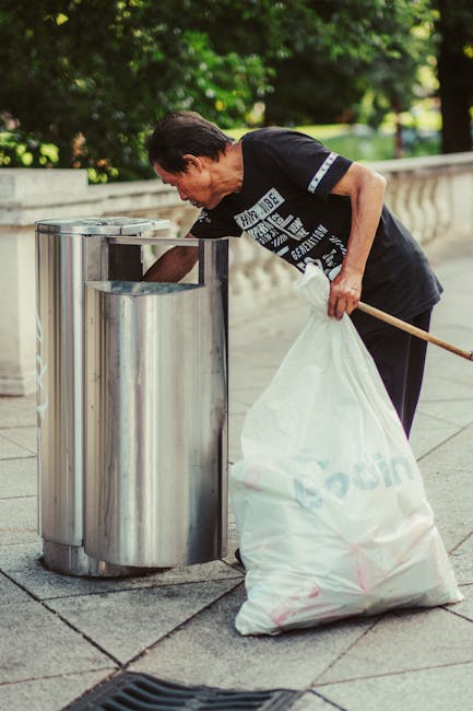 A middle-aged man with dark hair, wearing a black T-shirt with white graphic text and black trousers, is crouched slightly while depositing waste into a silver, cylindrical public litter bin situated on a paved area next to a stone balustrade. He holds a large, partially filled white rubbish bag with the word 'Recycling' faintly visible, using his left hand and supports it with his right hand resting on his knee. The bin has a sleek, reflective stainless steel surface with a rectangular opening at the top, and it is positioned outdoors amid greenery, including trees with lush leaves, suggesting a park or public promenade setting. The area is well-lit, and the scene emphasizes proper waste disposal, aligning with external rubbish removal or private waste handling services, as represented by Rubbish Collection Brixton.