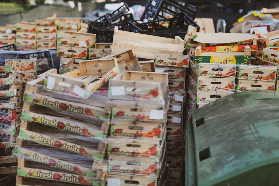 A large pile of empty wooden fruit crates, primarily made of light-colored, weathered wood with visible grain and a smooth finish, stacked irregularly outdoors on what appears to be a street or yard. Some crates display colorful labels with images of fresh fruits, such as tomatoes, apples, and oranges, along with the words 'fresh fruits' on the side. The crates vary in orientation, with some lying flat and others tilted or leaning against each other, creating a cluttered appearance. In the background, there are black plastic crates, loosely piled and stacked, indicating ongoing or past sorting or disposal activities. To the right, a curved, weathered green rubbish bin with a rough, slightly dirty surface is partially visible, suggesting the scene involves waste management or collection, possibly by Rubbish Collection Brixton, a company specialising in independent rubbish removal services. The lighting appears natural, highlighting textures and the muted colors of the materials, consistent with an outdoor environment prepared for waste collection or recycling activities.